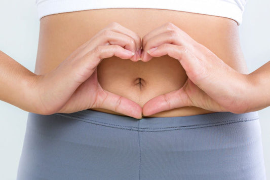 Woman with a white crop top on and blue yoga pants making a heart shape with her hands over her stomach