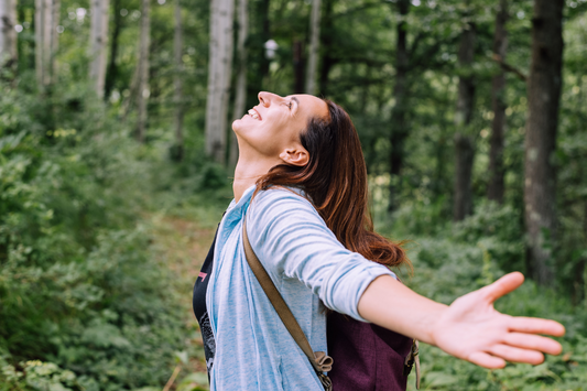 woman smiling with open arms in the woodlands with lots of greenery and trees, she is wearing a blue cardigan and a brown backpack