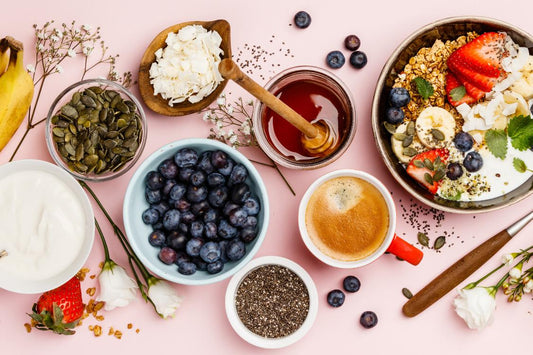 Breakfast food display with greek yoghurt, blueberries, chia seeds, pumpkin sees, honey, yoghurt granola bowl with banana, strawberries, nuts and seeds on a pink background with flowers