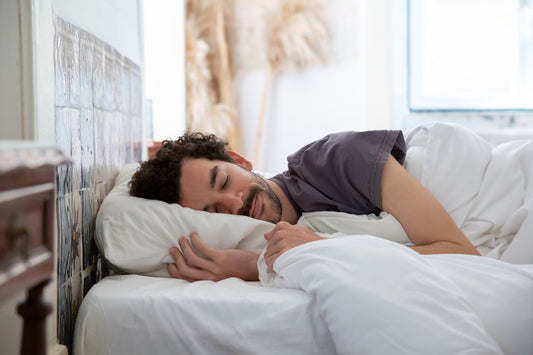 A bearded man peacefully sleeping with a dark grey t-shirt in a white linen-covered bed in a bedroom.