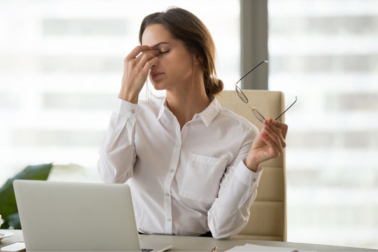 Woman in a white blouse sat in front of laptop holding her glasses in one hand and pressing her fingers onto her nose in stress and fatigue