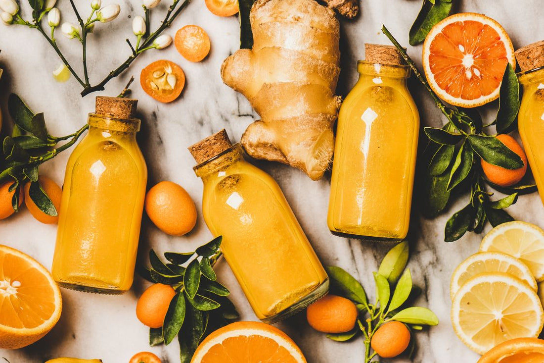 three bottles of lemon, orange, and ginger juice, surrounded by sliced oranges, ginger roots, and green leaves