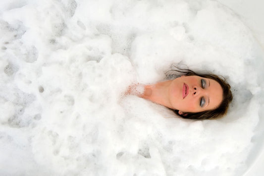 woman relaxing in a bubble bath with her eyes closed with brown hair, white skin and wearing facial make up
