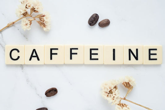 Wooden Scrabble letters spell 'caffeine' on a marble background surrounded by coffee beans and coffee flowers, symbolizing the essence of this energizing molecule amidst natural coffee elements.