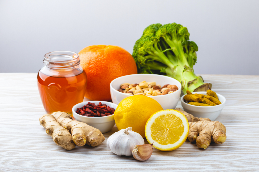 immune boosting foods on a white and grey table background, honey, orange, lemon, broccoli, garlic, ginger, nuts and seeds