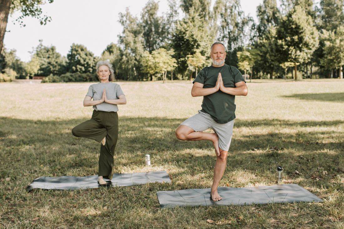 an older couple with grey hair doing yoga in a green park with grey yoga matts in the summer