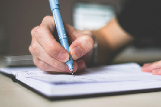 white hand holding a blue pen making notes in a black and white diary, wearing a black t-shirt