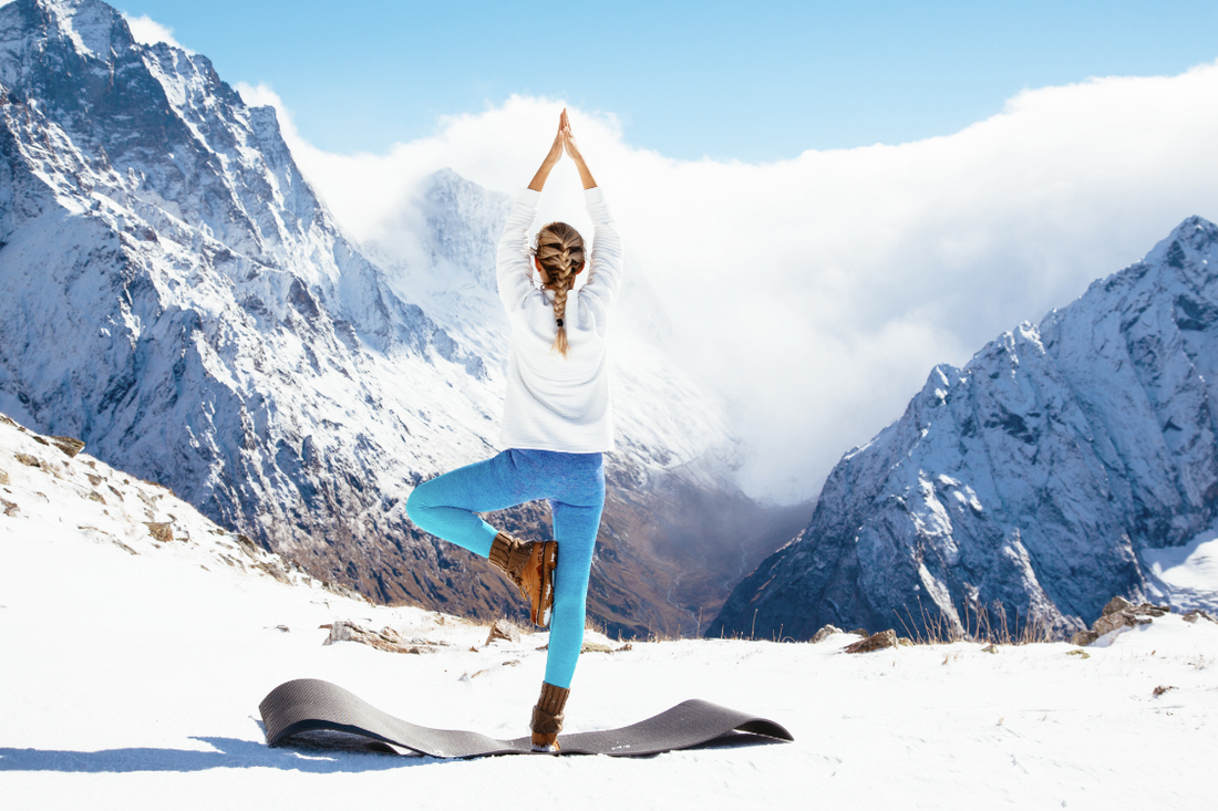 woman doing a yoga pose in a blue skies snowy mountain with blonde plaited hair, brown boots, blue trousers, and white jumper