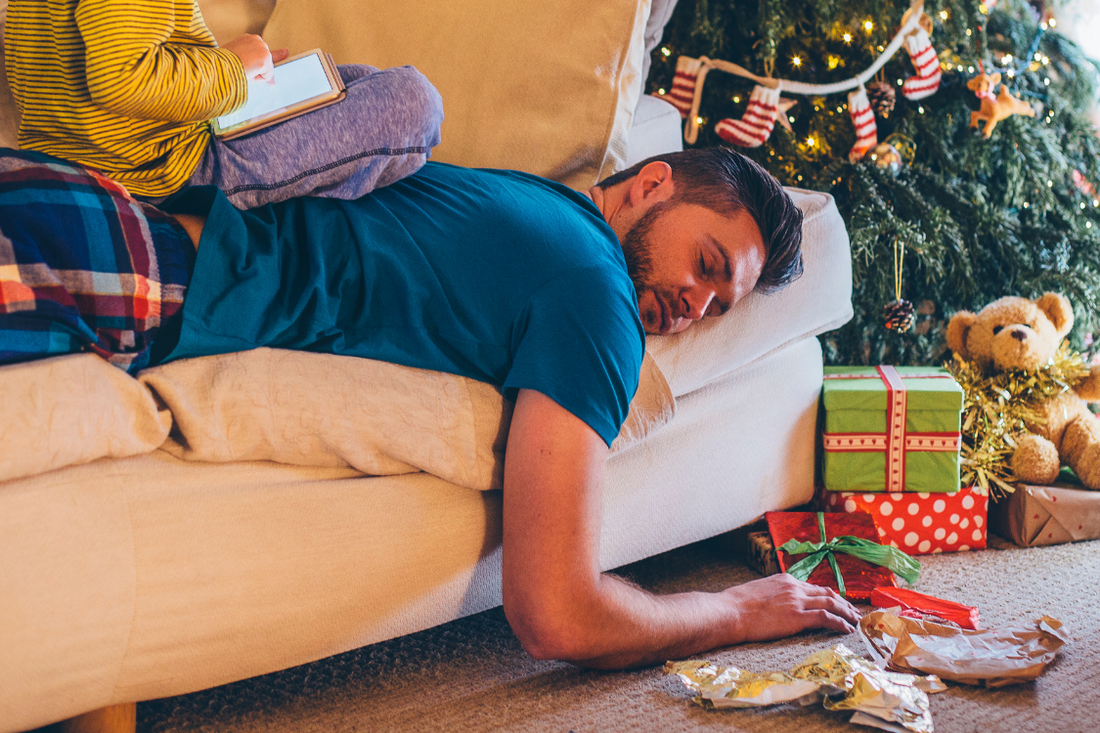 Exhausted man lying on a white couch at Christmas, with a child on his back scrolling on a phone and a christmas tree in the background