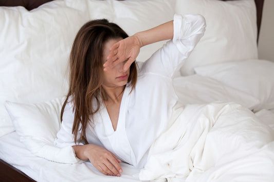 Woman wearing a white pyjama shirt, waking up in a white bed, looking uncomfortable due to the heat