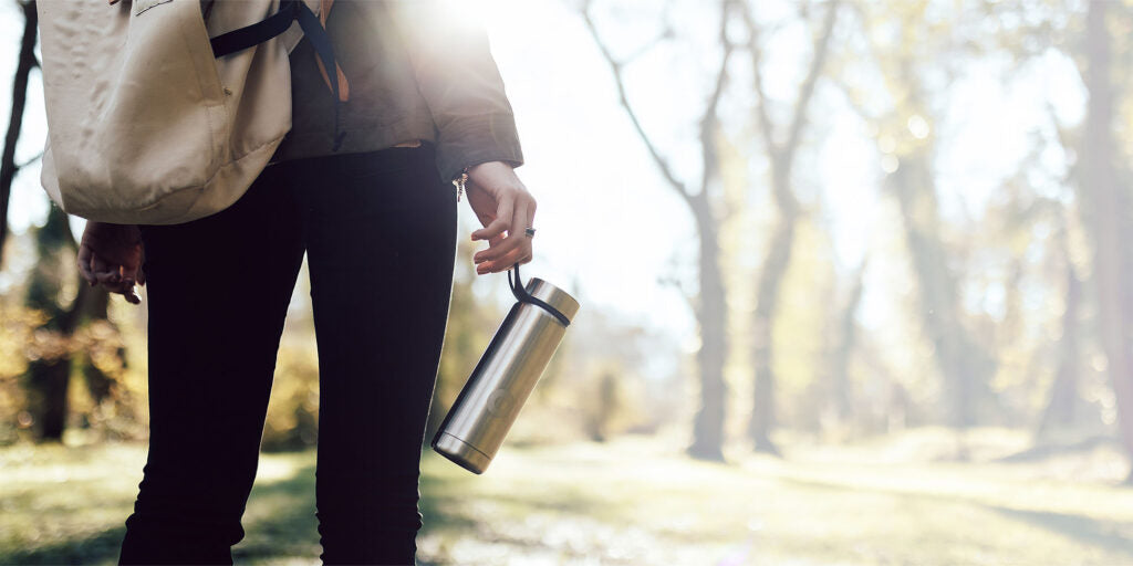 Woman walking in the sunny woods with black trousers holding a silver insulated bottle and carrying a backpack